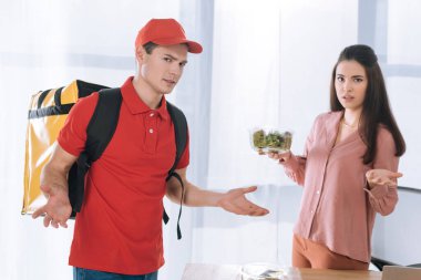 Kafası karışmış kurye, iş kadınının yanındaki kameraya bakıyor. Ofisinde salata var.