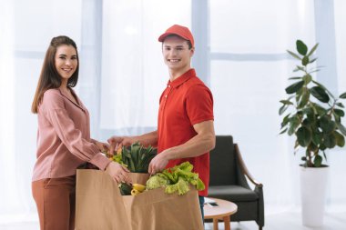 Side view of courier smiling at camera and giving packages with fresh vegetables to attractive woman  clipart