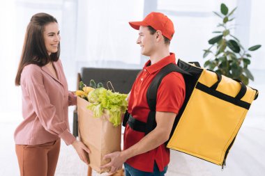 Side view of courier with thermo backpack giving package with fresh vegetables to smiling woman at home clipart