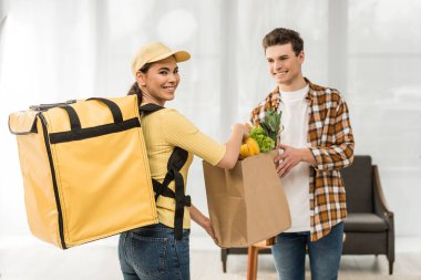 Beautiful courier smiling at camera and giving package with fresh vegetables to man at home clipart