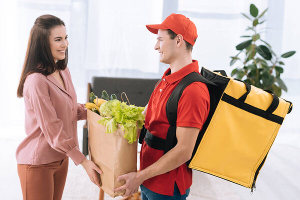 Side view of courier with thermo backpack giving package with fresh vegetables to smiling woman at home