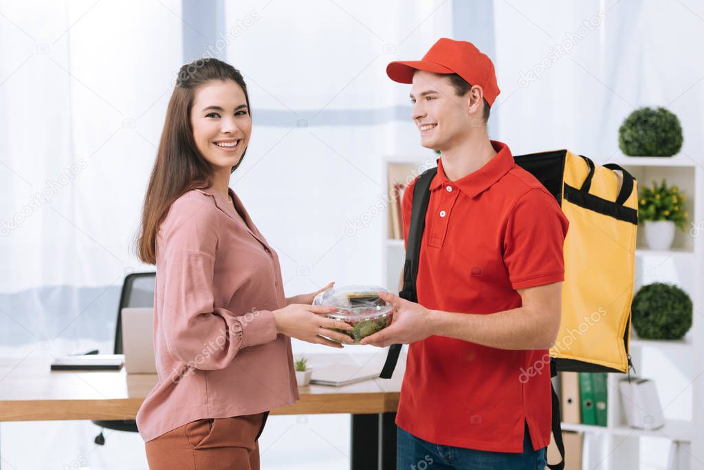 Businesswoman smiling at camera while holding container with salad near delivery man with thermo backpack in office