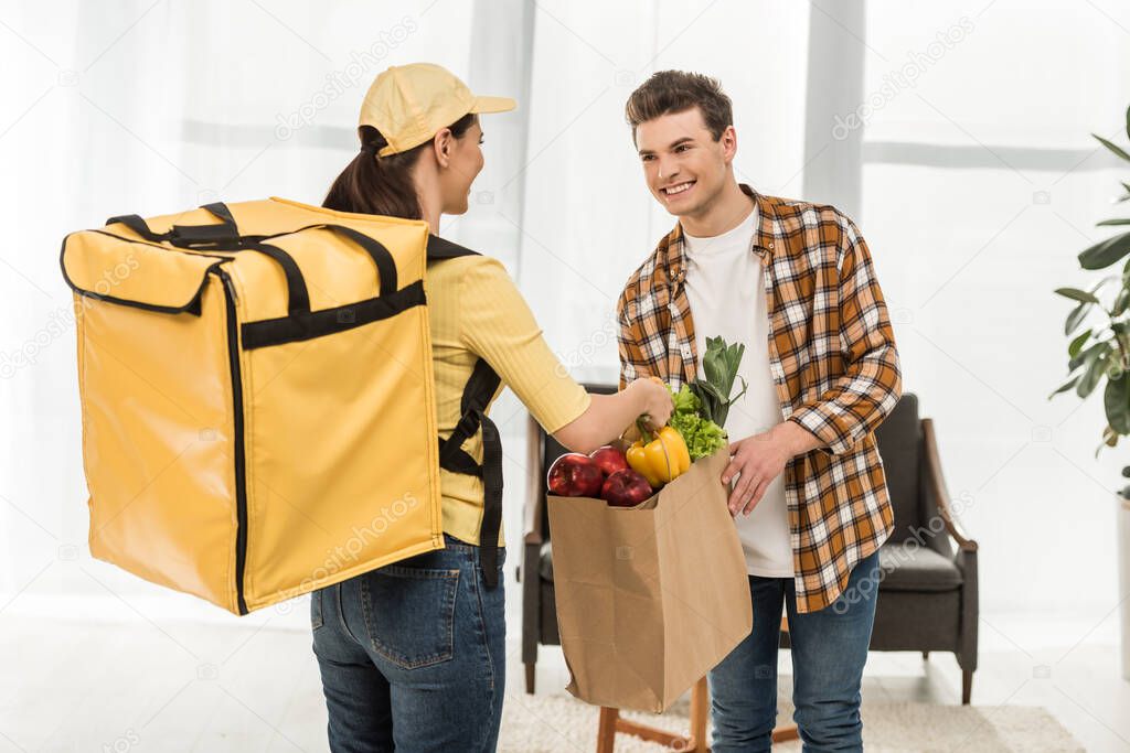 Courier giving package with fresh vegetables to smiling man at home 