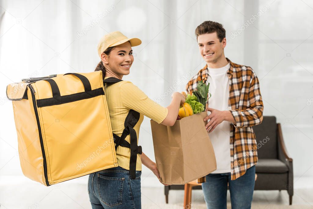Beautiful courier smiling at camera and giving package with fresh vegetables to man at home