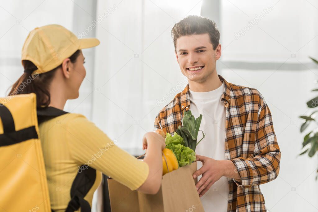 Selective focus of courier in yellow uniform giving package with fresh vegetables to smiling man 