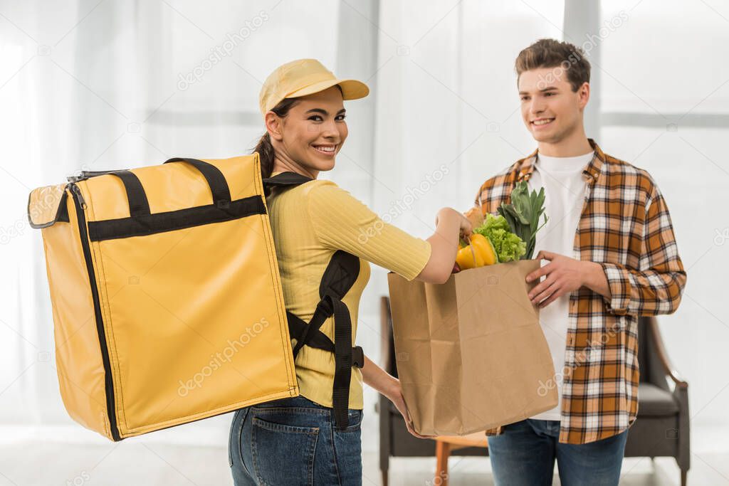 Attractive courier smiling at camera and giving package with fresh vegetables to man at home
