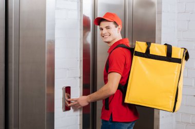 Side view of courier with thermo backpack smiling at camera and pressing elevator button  clipart