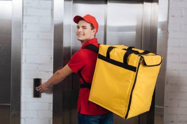 Delivery man with thermo backpack smiling at camera and pressing elevator button clipart