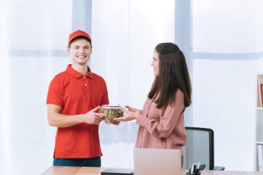 Gülümseyen teslimatçı masadaki iş kadınına paket salata veriyor.
