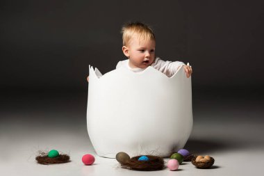 Child with open mouth holdind eggshell while sitting inside on black background clipart