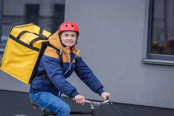 Delivery man with thermo backpack smiling at camera and riding bicycle near building