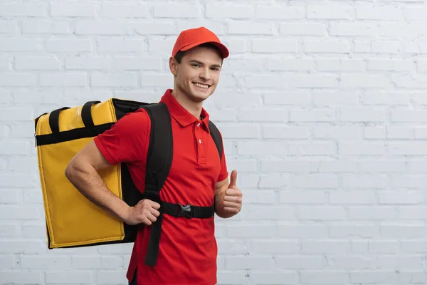 Smiling delivery man with thermo backpack smiling at camera and showing thumb up near brick wall