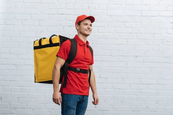 Handsome delivery man with thermo backpack smiling away near brick wall