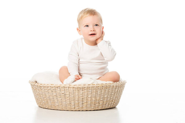 Child touching neck and looking up, sitting on blanket in basket on white background
