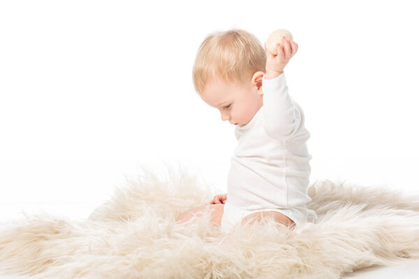 Side view of cute kid looking down and holding easter egg in raised hand, sitting on fur on white background