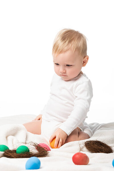 Симпатичный ребенок держит пасхальное яйцо, глядя вниз, сидя на одеяло изолированы на белом
