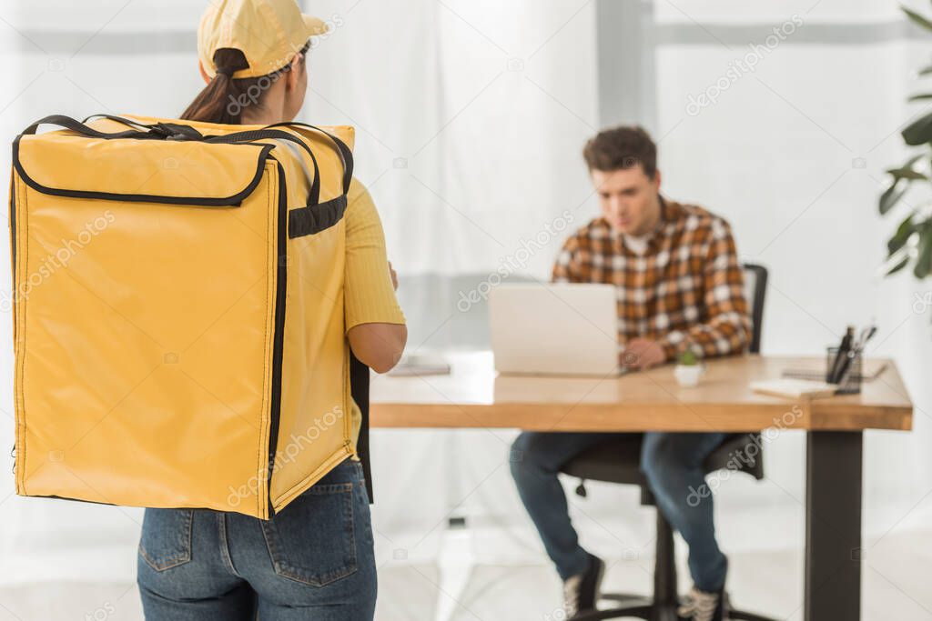 Selective focus of courier with thermo backpack and businessman using laptop at table