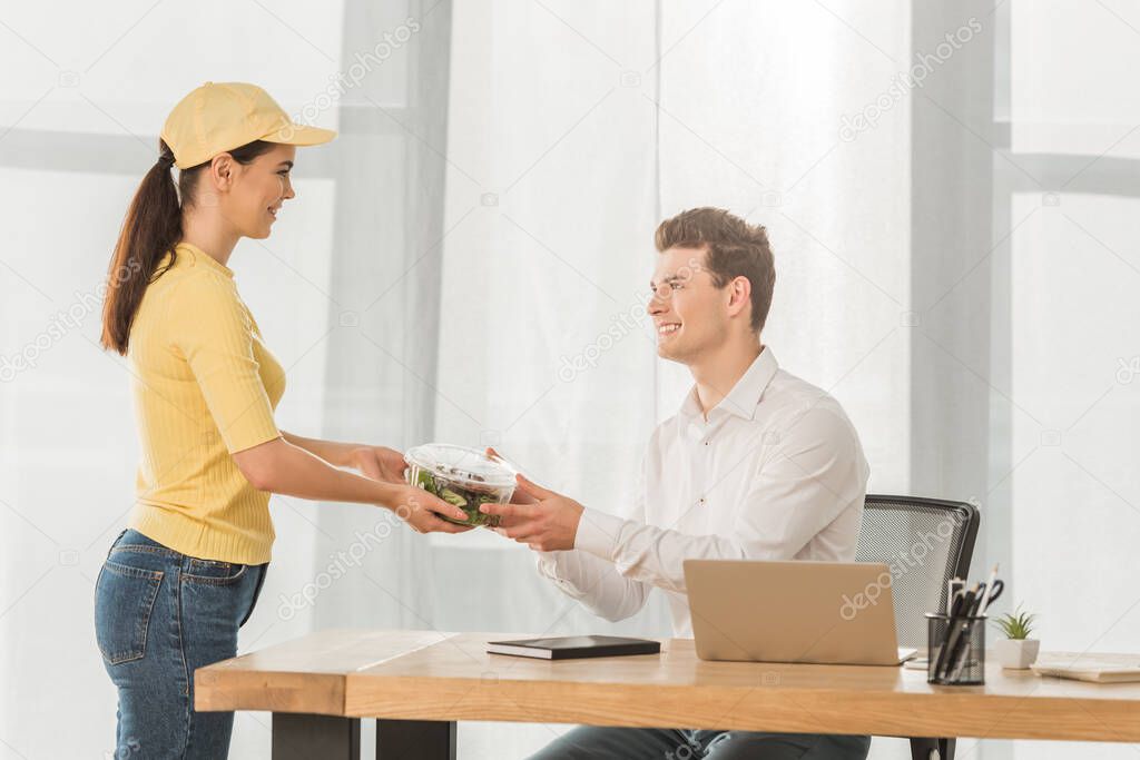 Side view of smiling courier giving container with salad to businessman at table
