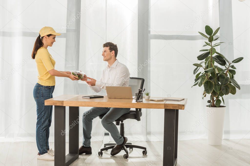 Side view of courier giving takeaway salad to smiling businessman at table in office