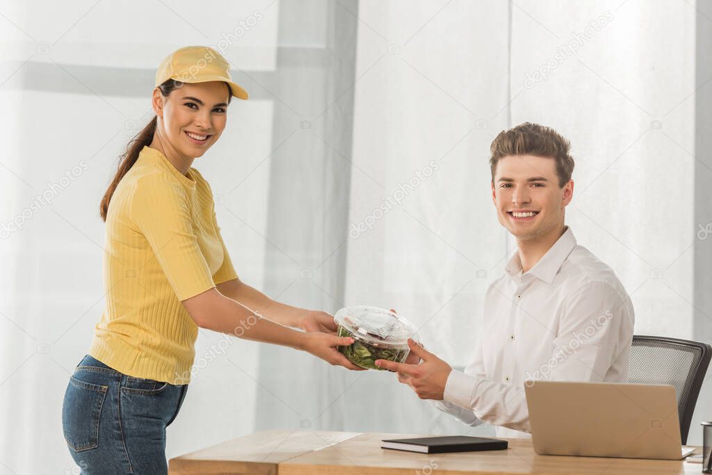 Side view of courier giving salad to businessman and smiling at camera in office