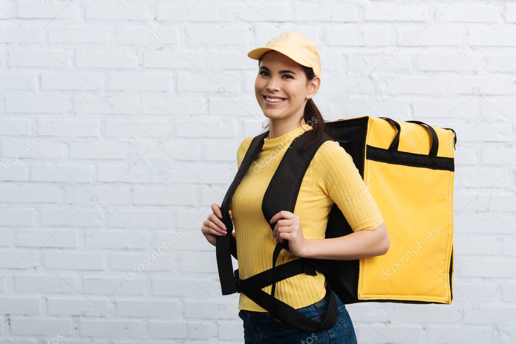 Beautiful courier in yellow uniform carrying thermo backpack and smiling at camera near brick wall