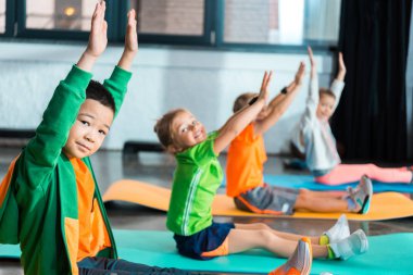 Çok ırklı çocukların seçmeli odak noktası kameraya elleri havada bakmak ve spor salonundaki spor paspasları üzerinde çalışmak.