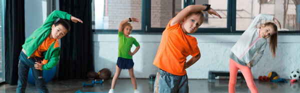 Selective focus of multicultural children warming up on fitness mats in gym, panoramic shot
