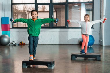 Çok kültürlü çocuklar ellerini uzatıp dambılları tutuyorlar ve spor salonunda ayaklı platformlarda egzersiz yapıyorlar.