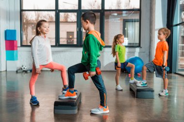Selective focus of multiethnic children holding dumbbells and working out together on step platforms in gym clipart