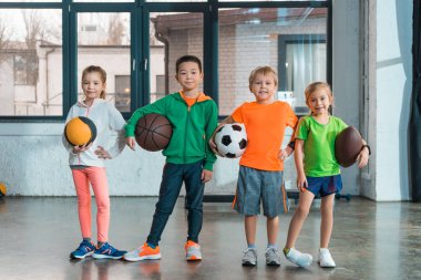 Front view of multiethnic children smiling and holding balls in gym clipart