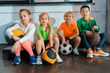 Çok kültürlü çocuklar spor salonunda toplarıyla merdivenlerde oturuyorlar.