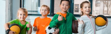 Front view of happy multicultural children holding balls in gym, panoramic shot clipart