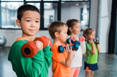 Çok ırklı çocukların seçici odak noktası, ellerinde halterlerle spor salonunda.