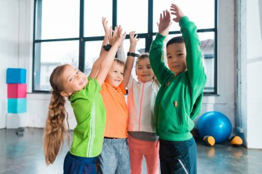 Multiethnic children raising hands up together in gym clipart