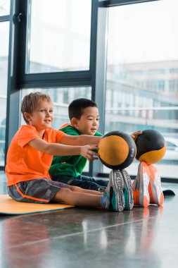 Çok kültürlü çocuklar ayak parmaklarının ucuna top koyup spor salonundaki spor paspasına oturuyorlar.