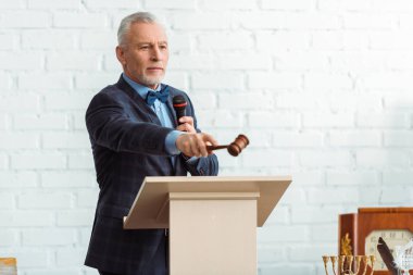 Takım elbiseli yakışıklı bir müzayedeci mikrofon tutuyor ve açık arttırma sırasında tokmakla işaret ediyor.