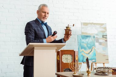 Takım elbiseli yakışıklı bir müzayedeci elinde kahve öğütücüsüyle açık arttırma sırasında mikrofonu tutuyor.