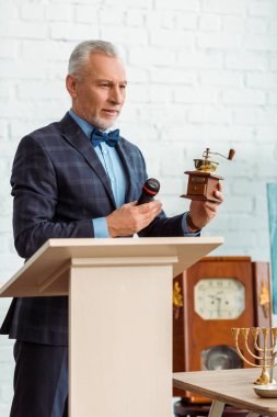 Takım elbiseli yakışıklı bir müzayedeci elinde kahve öğütücüsüyle açık arttırma sırasında mikrofonu tutuyor.