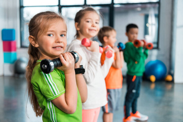 Selective focus of multiethnic children lined up, holding dumbbells and smiling in sports center