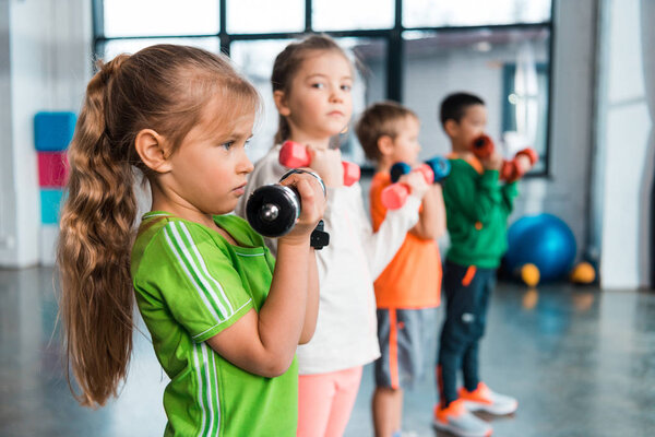 Selective focus of multicultural children lined up, holding dumbbells in sports center