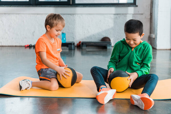 Selective focus of multicultural kids looking at balls in hands and sitting on fitness mats in gym