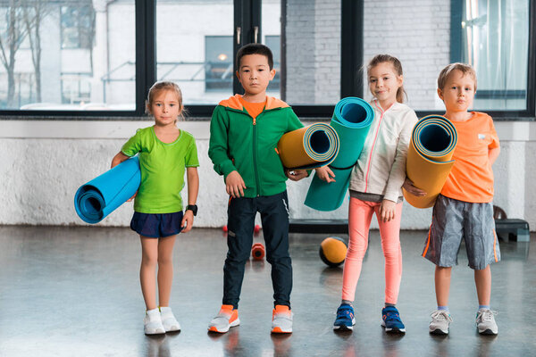 Front View of multicultural children holding fitness mats and looking at camera in gym