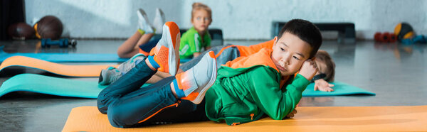 Selective focus of multicultural children lying on fitness mats in sports center, panoramic shot