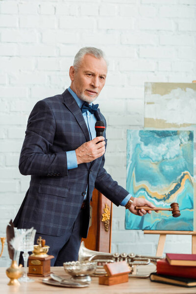 аукционист в костюме разговаривает с микрофоном и держит молоток во время аукциона
