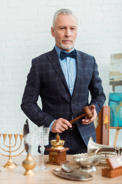 красивый аукционист в костюме держит молоток и смотрит в камеру во время аукциона
