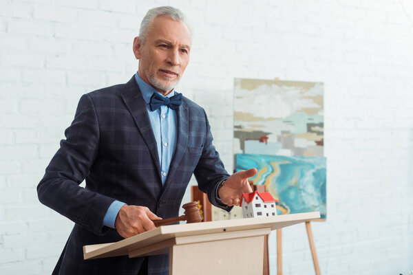 аукционист, указывающий рукой на модель дома и держащий молоток во время аукциона

