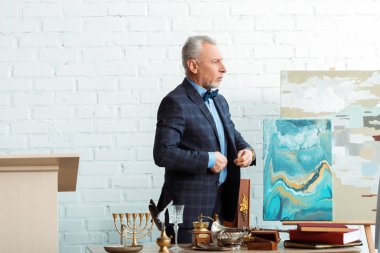 Eski nesnelerin yanında yakışıklı ve olgun bir müzayedeci ceketi ilikliyor. 