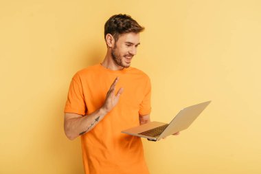 cheerful young man waving hand while having video call on laptop on yellow background clipart