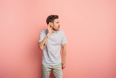 confused young man showing cant hear gesture by holding hand near ear on pink background clipart