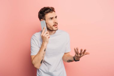 shocked young man standing with open arm while talking on smartphone on pink background clipart